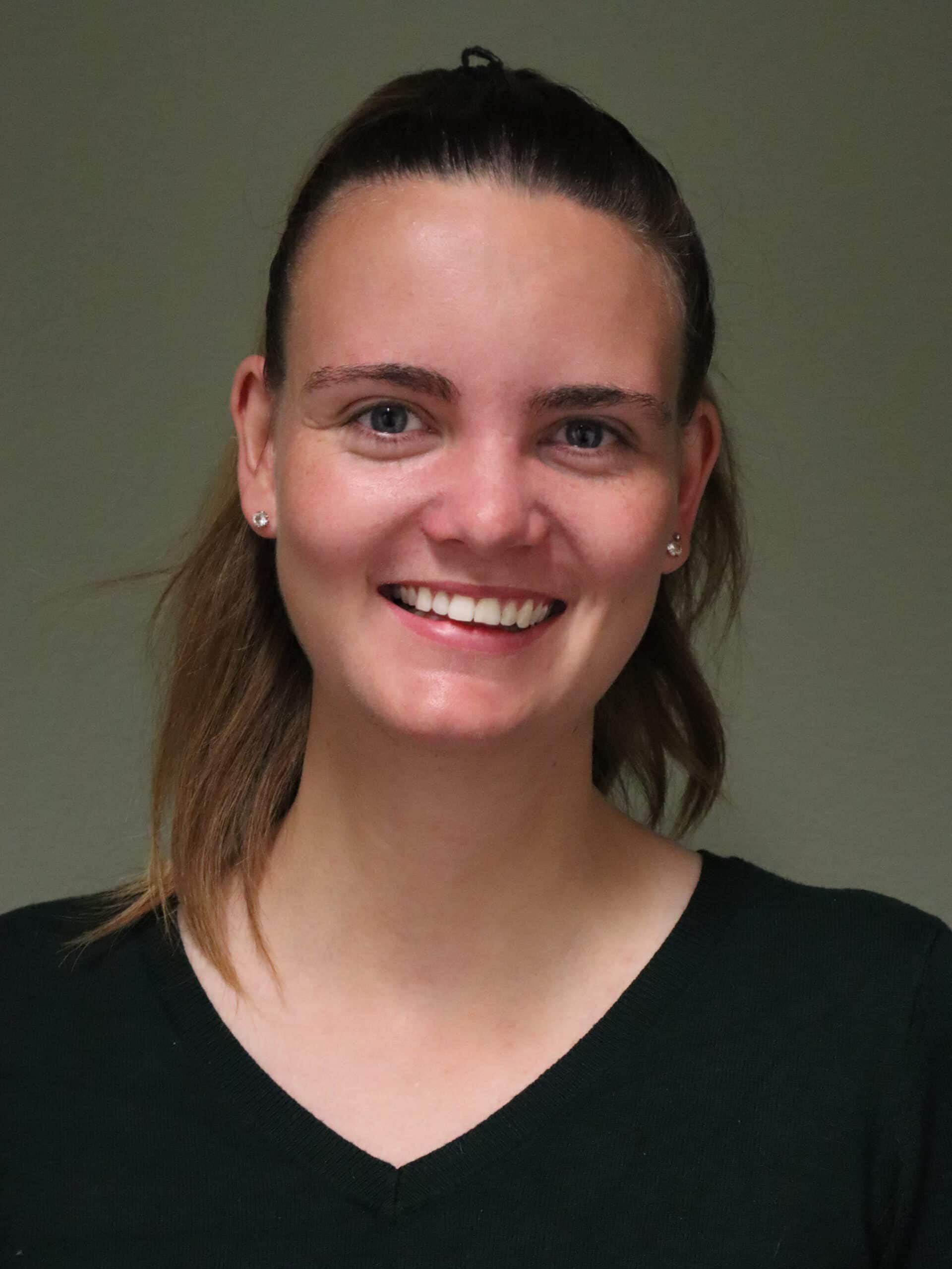 Lächelnde Frau mit braunen Haaren und Ohrringen vor neutralem Hintergrund.