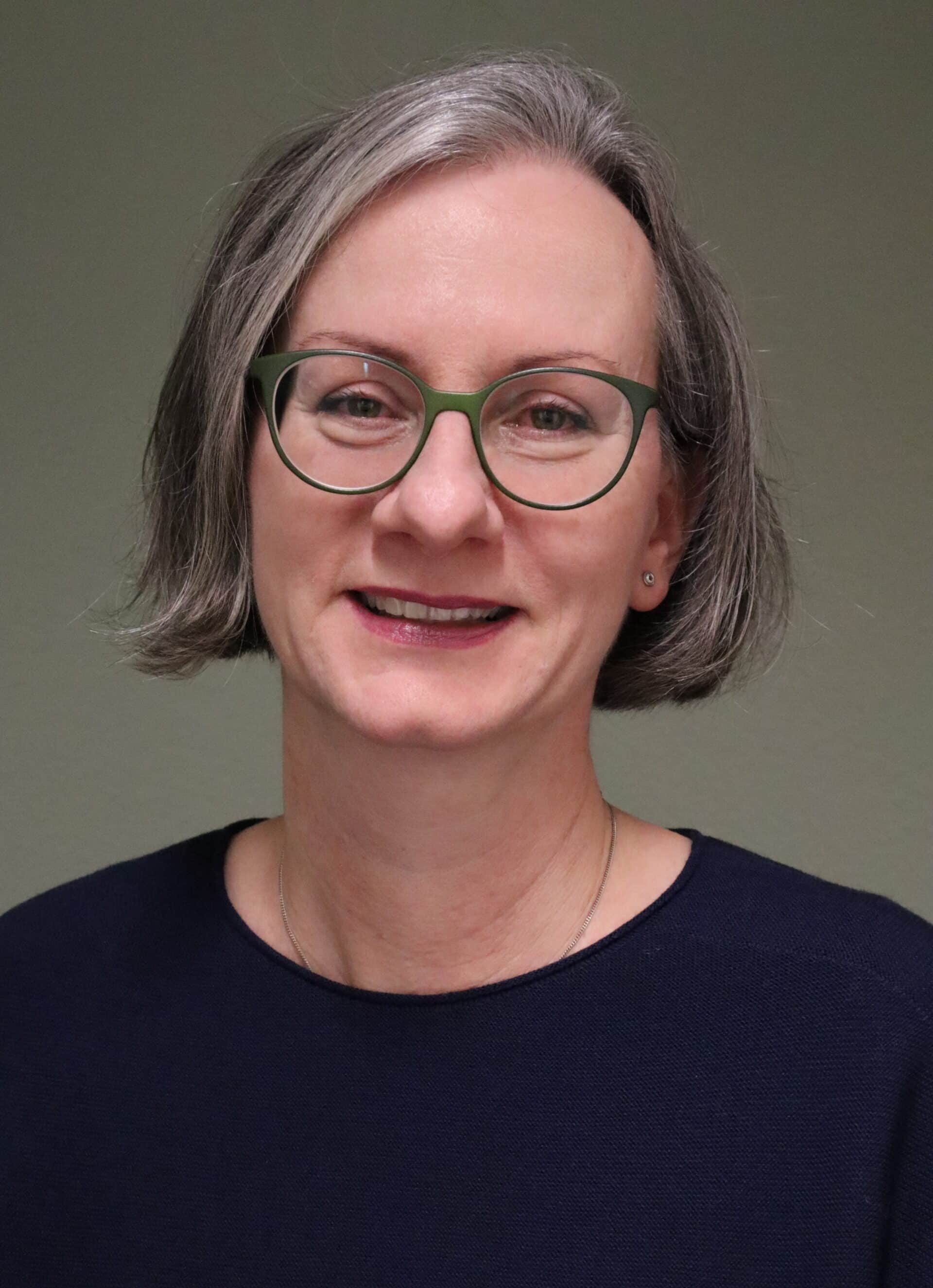 Eine lächelnde Frau mit Brille und grauen Haaren vor neutralem Hintergrund.