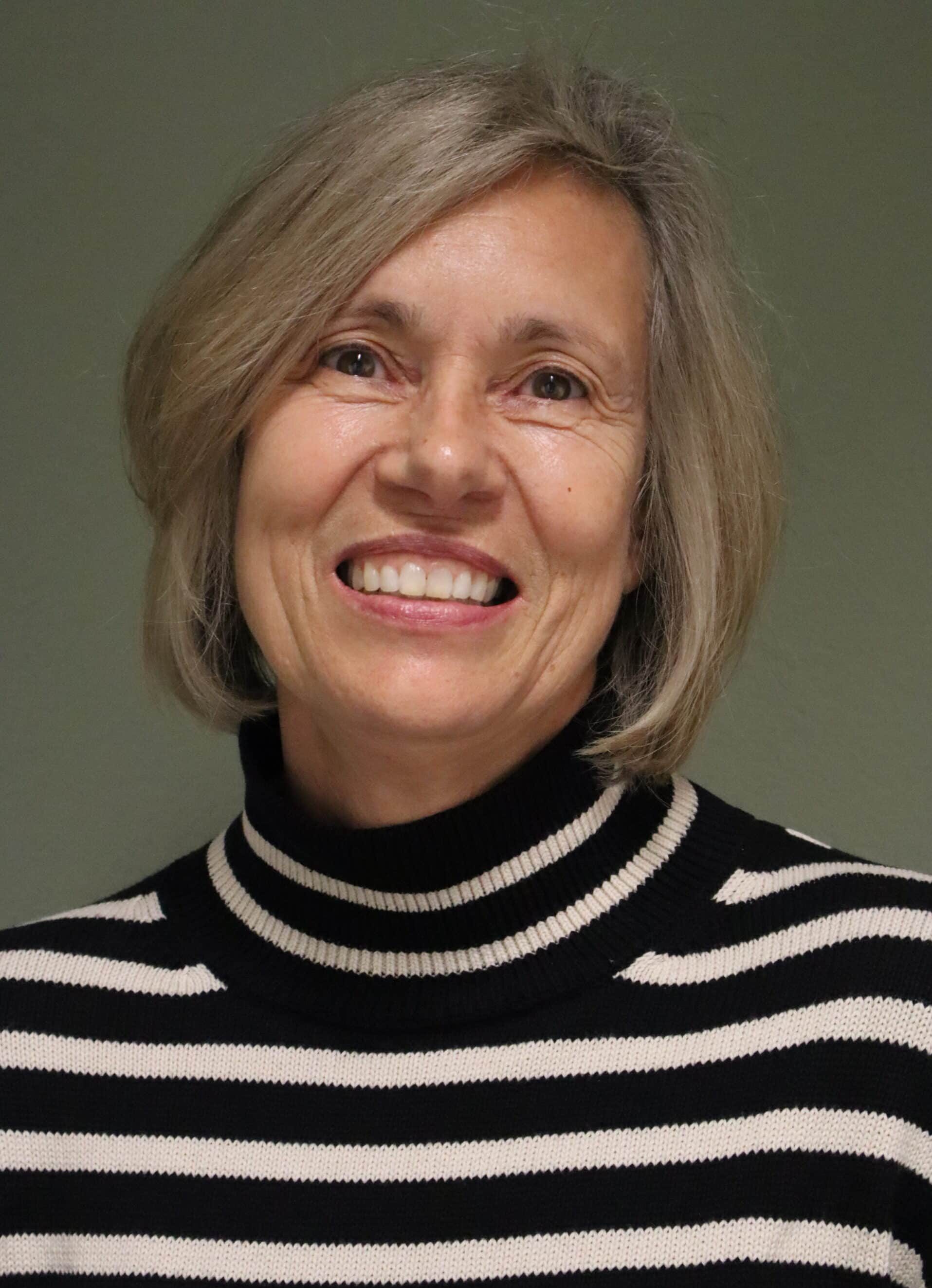 Frau mit kurzen grauen Haaren lächelt freundlich, trägt einen schwarz-weiß gestreiften Pullover vor neutralem Hintergrund.