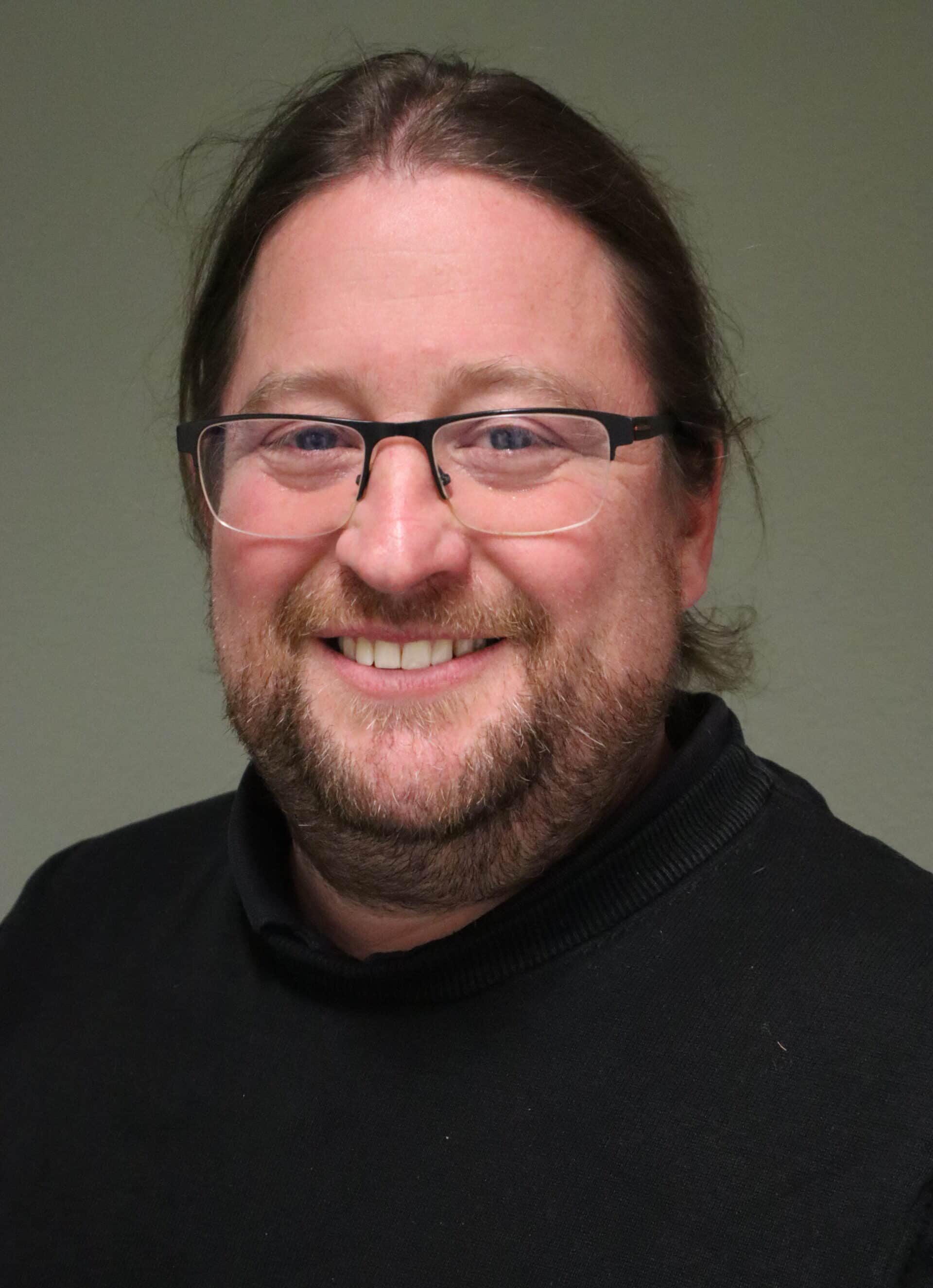 Lächelnder Mann mit Brille und dunklem Pullover vor neutralem Hintergrund.