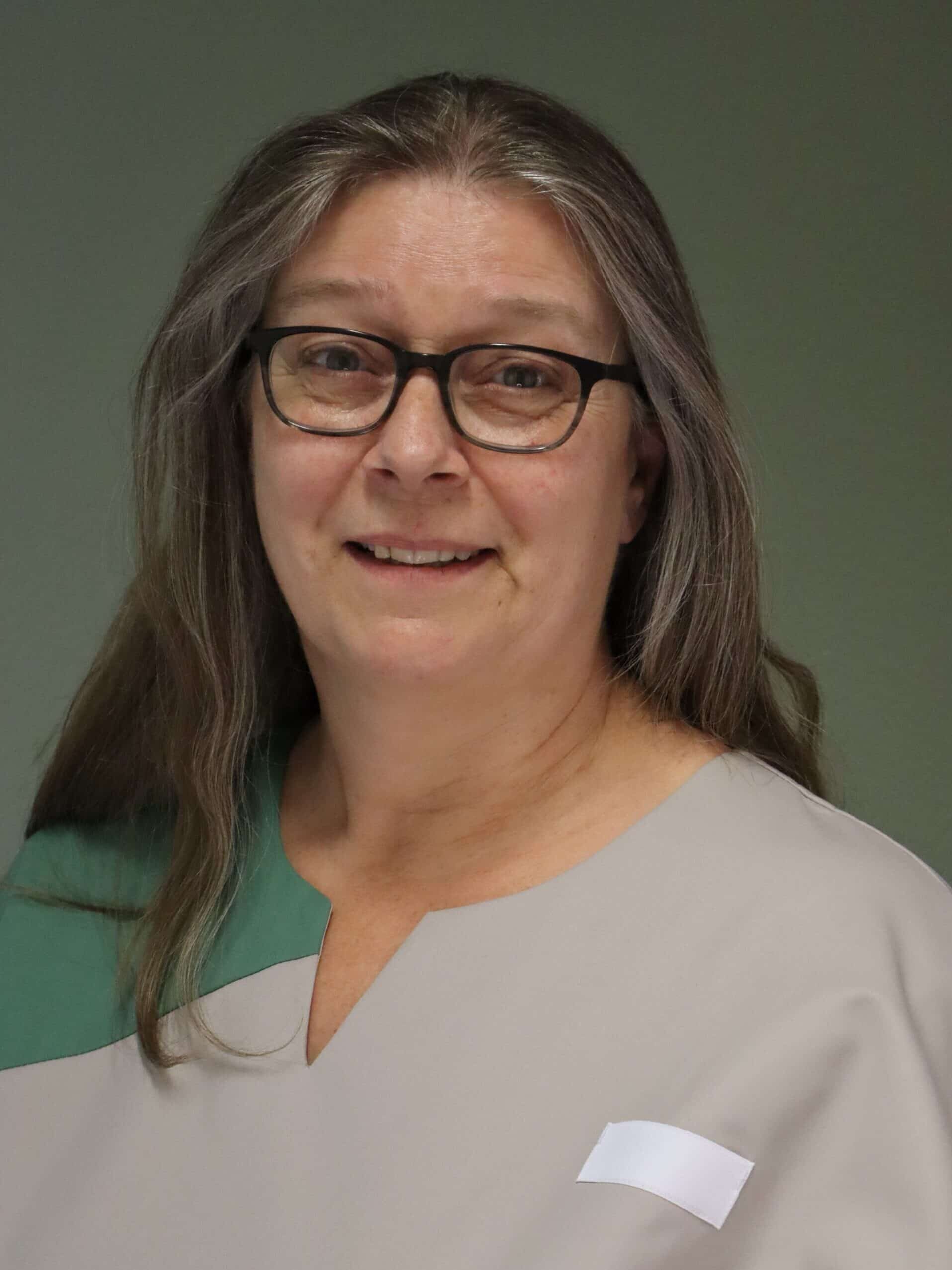 Ältere Frau mit Brille und langen Haaren in grün-beiger Kleidung, lächelnd vor neutralem Hintergrund.