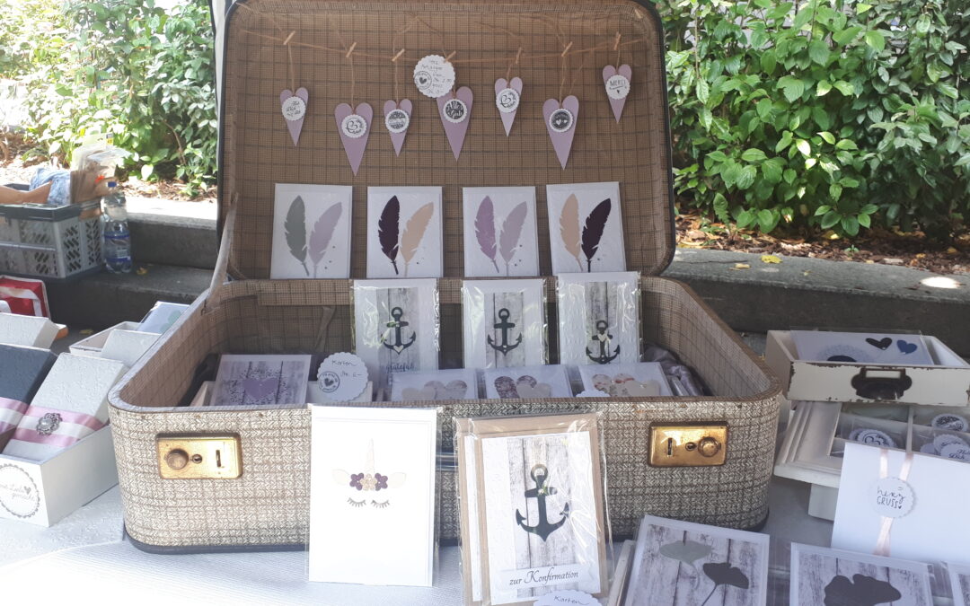 Offener Koffer mit handgefertigten Grußkarten und Dekorationen auf einem Marktstand, umgeben von Pflanzen.