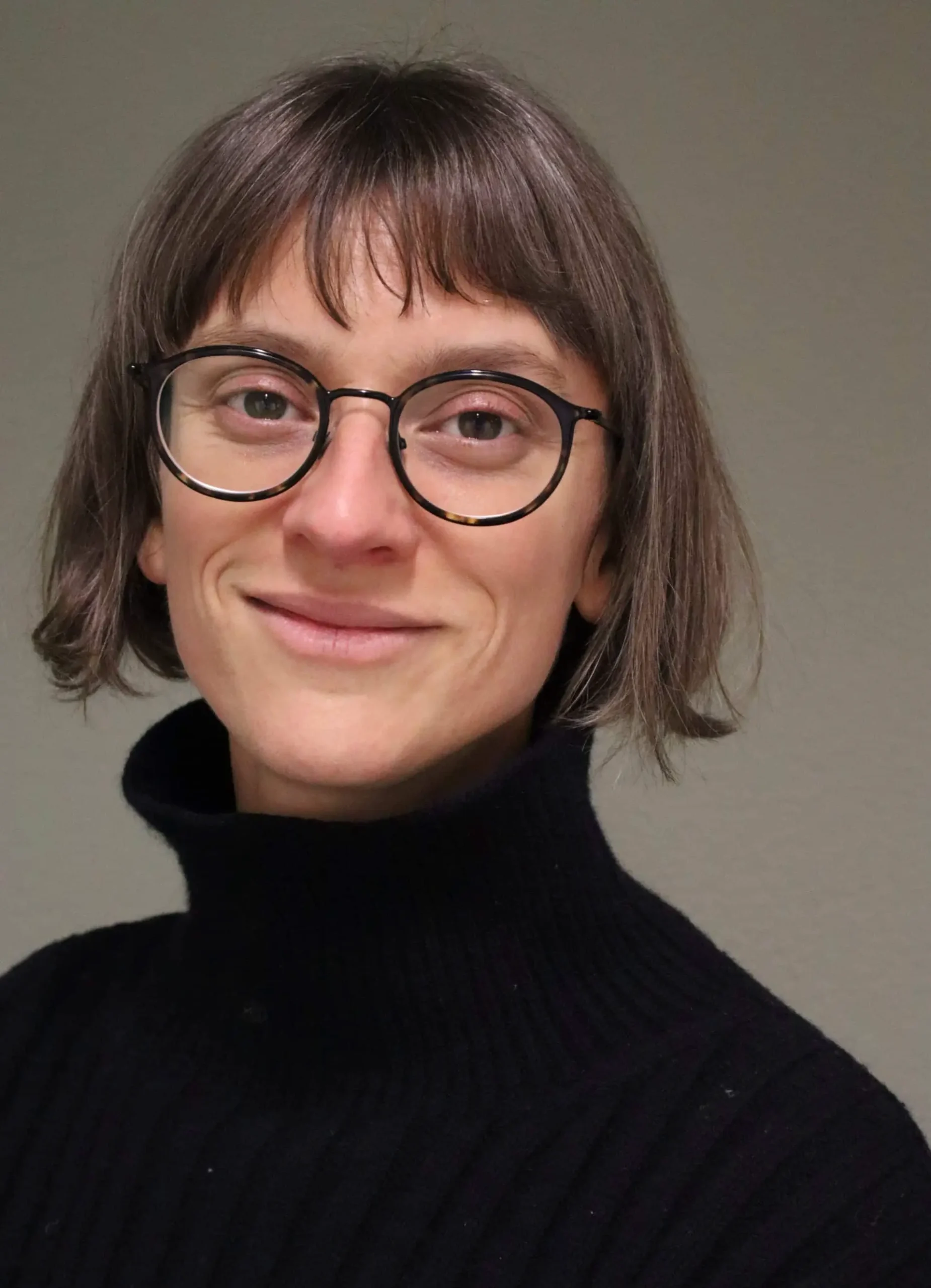 Porträt einer Person mit Brille und schwarzem Rollkragenpullover, lächelnd vor neutralem Hintergrund.